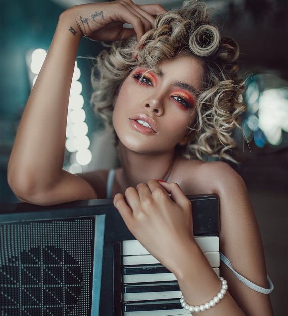 Self assured young female singer with curly hair and makeup standing in modern studio with synthesizer before concert